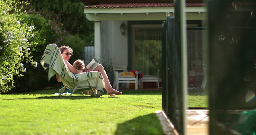 A young woman and child are seated in the backyard of a suburban under the warm sunshine and surrounded by a lush lawn. 