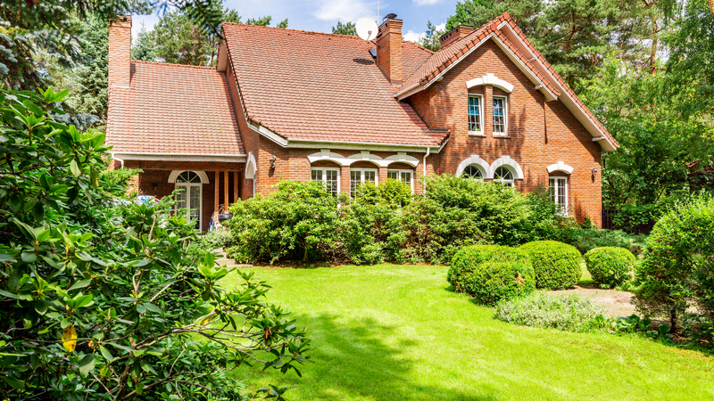 A large suburban home looms over a lush green lawn and surrounding shrubs. 